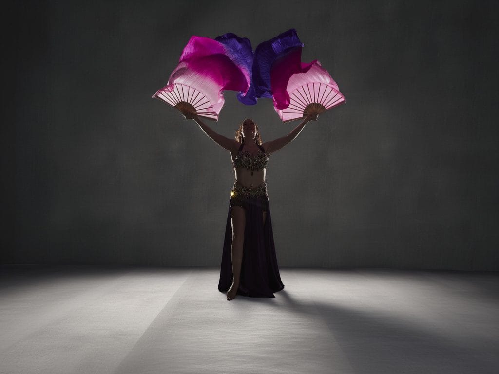Aurore, danseuse orientale, pose avec deux grands éventails en soie fuchsia et violet dans un studio sombre avec un éclairage dramatique.
