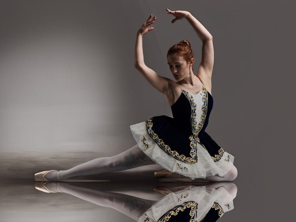 La danseuse Eloria, en costume de ballet bleu marine, blanc et doré, réalise une pose de grand écart au sol. Ses bras sont levés au-dessus de sa tête. Son reflet est visible sur le sol brillant du studio.