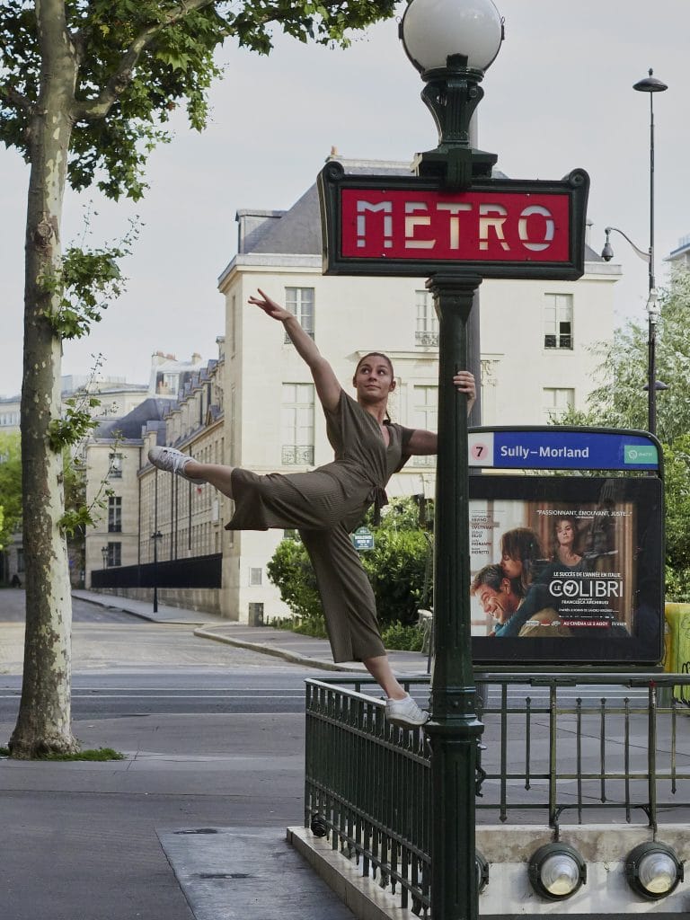 La danseuse Clara, en combinaison kaki et baskets, s'équilibre sur une rambarde d'escalier de métro à Paris. Elle tient le poteau du panneau Métro de la station Sully-Morland, la jambe gauche levée, dans une pose de danse.