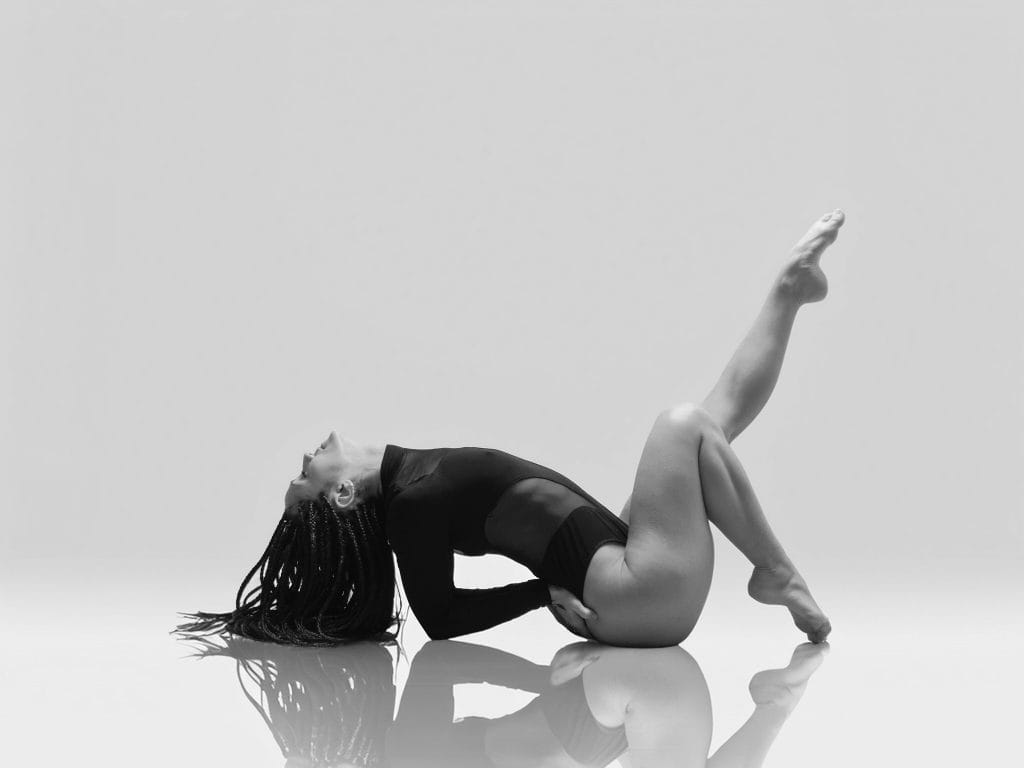 Wayna, danseuse, effectue une contorsion sur le dos, en body noir, sur un sol réfléchissant de studio, en noir et blanc.