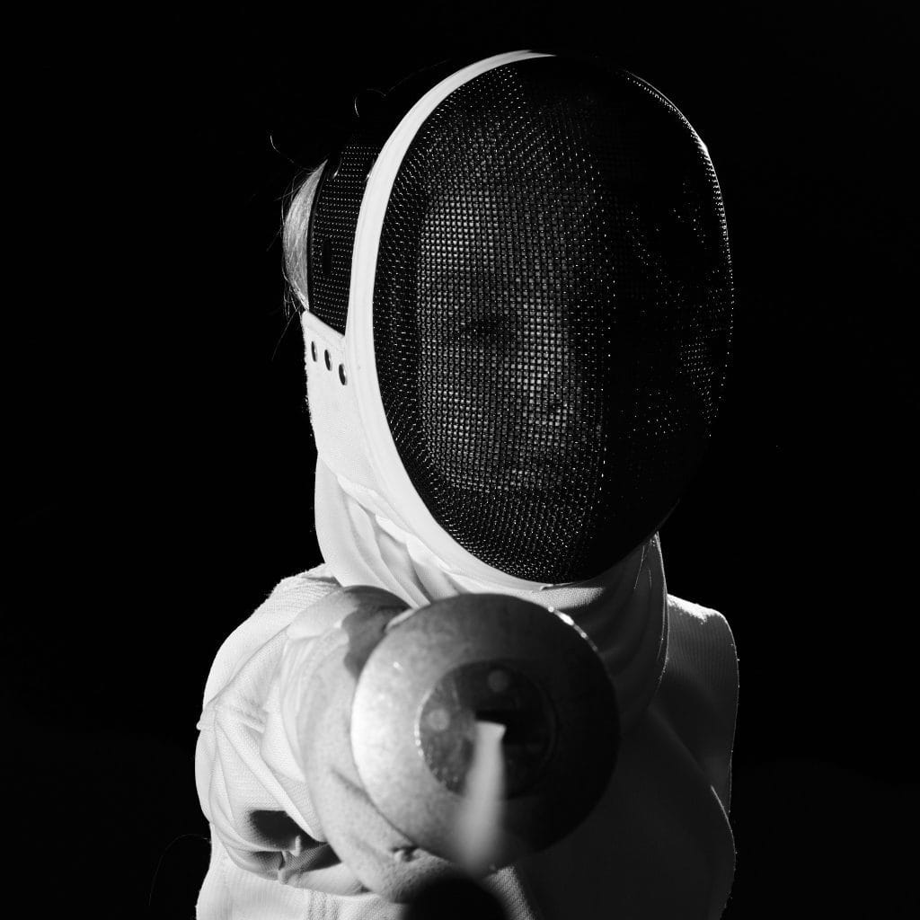 Gros plan d'un escrimeur en noir et blanc, avec son masque et son épée. Il regarde droit vers l'objectif, l'épée pointant vers la caméra.