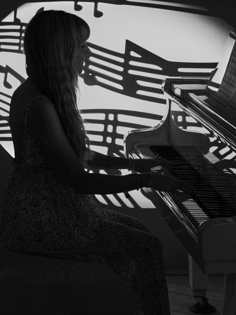 Femme de profil, Nelly joue du piano à queue, avec des ombres de notes de musique projetées derrière elle. Photo en noir et blanc.