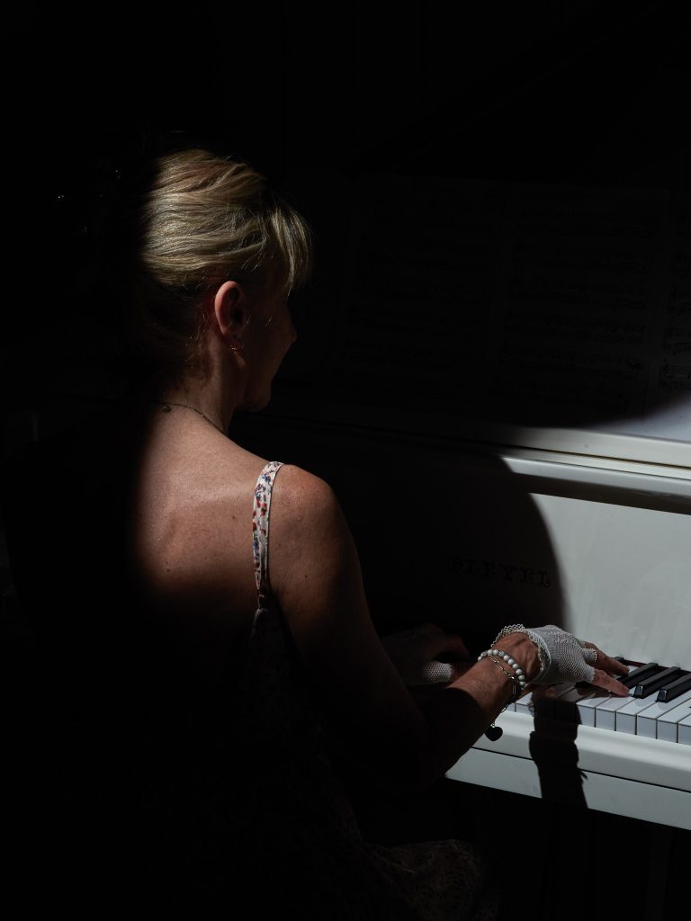 Vue de dos de Nelly, une femme aux cheveux blonds, jouant intensément du piano à queue Pleyel blanc. La lumière ne frappe que son épaule et ses mains gantées de blanc, créant une ambiance dramatique et intime, accentuant la concentration de l'artiste sur sa musique.