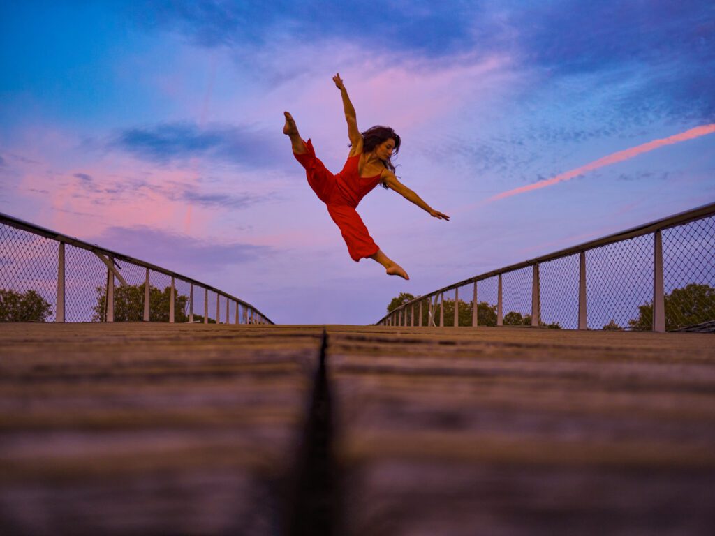 Clara, danseuse en combinaison rouge, exécute un grand saut artistique sur un pont en bois, sous un ciel de coucher de soleil à Paris sur la passerelle Simone de Beauvoir.