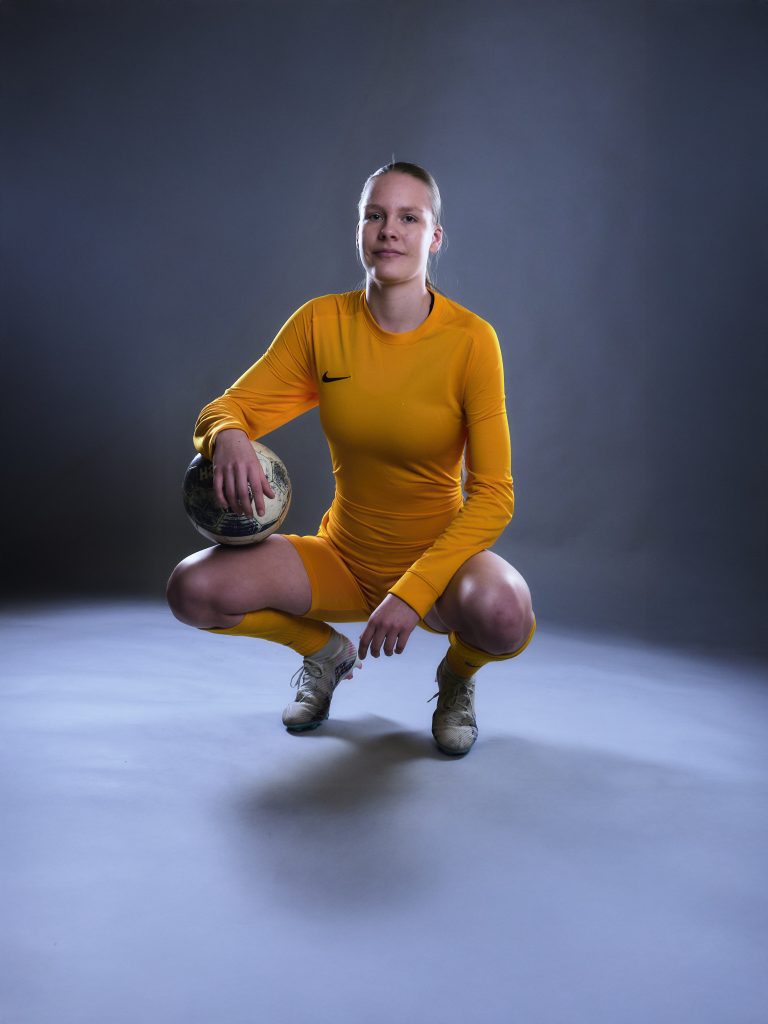 Wiktoria, gardienne de foot, en maillot, short et chaussettes jaunes, accroupie avec le ballon sous un bras, en appui sur une jambe.