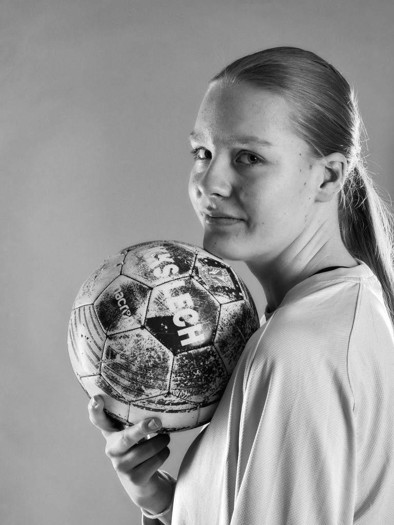 Portrait en noir et blanc de Wiktoria, une gardienne de but. Elle est de profil, regarde l'objectif, et tient un ballon de football sous son menton.