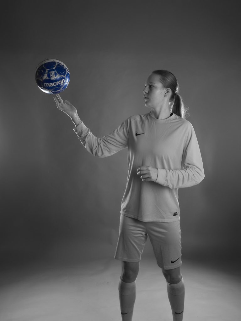 Portrait en noir et blanc de Wiktoria, une gardienne de but, jonglant avec un ballon de football bleu sur son doigt à hauteur de tête. Le ballon est le seul élément de couleur de l'image.