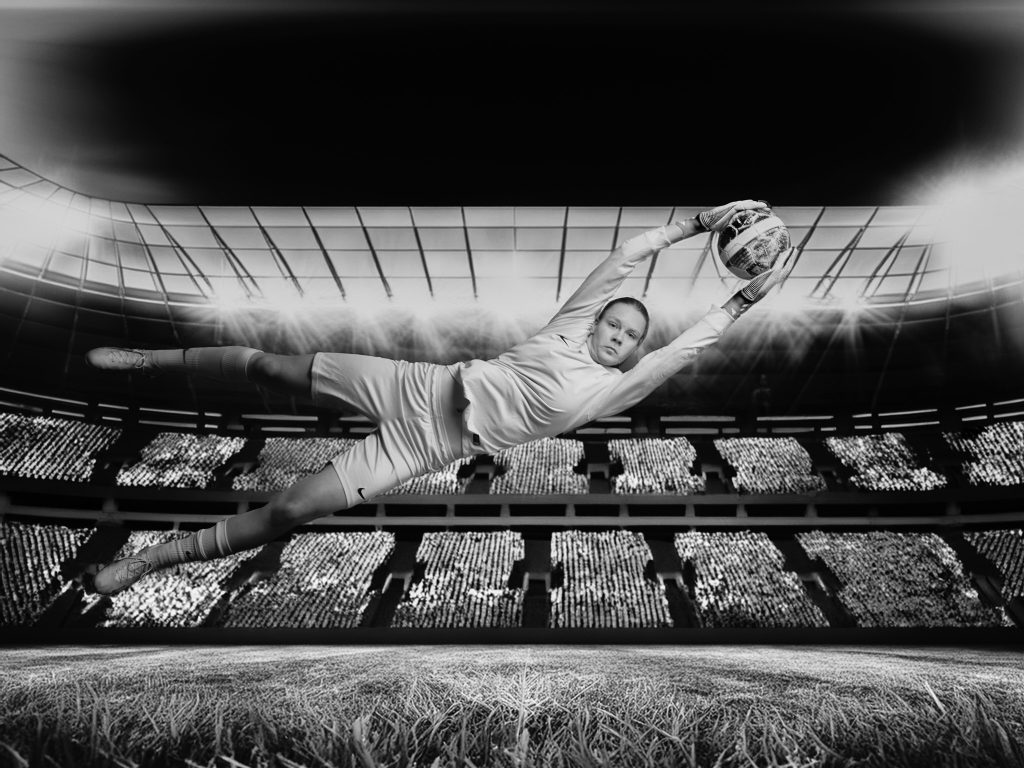 Wiktoria, gardienne de football, en noir et blanc, captée en plein saut latéral en studio avec un arrière-plan de stade de football.