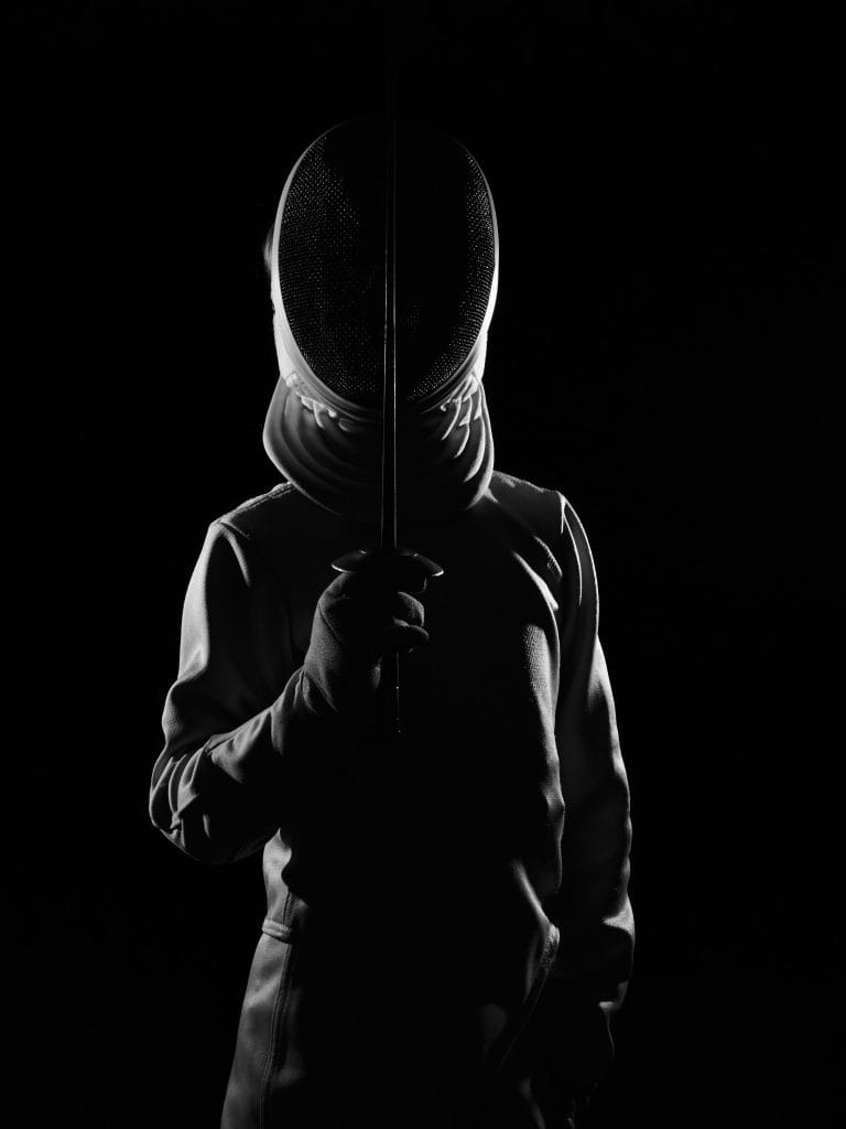 Photo en noir et blanc d'un escrimeur en tenue. Il tient son épée à la verticale, devant son masque, le visage dans l'ombre.