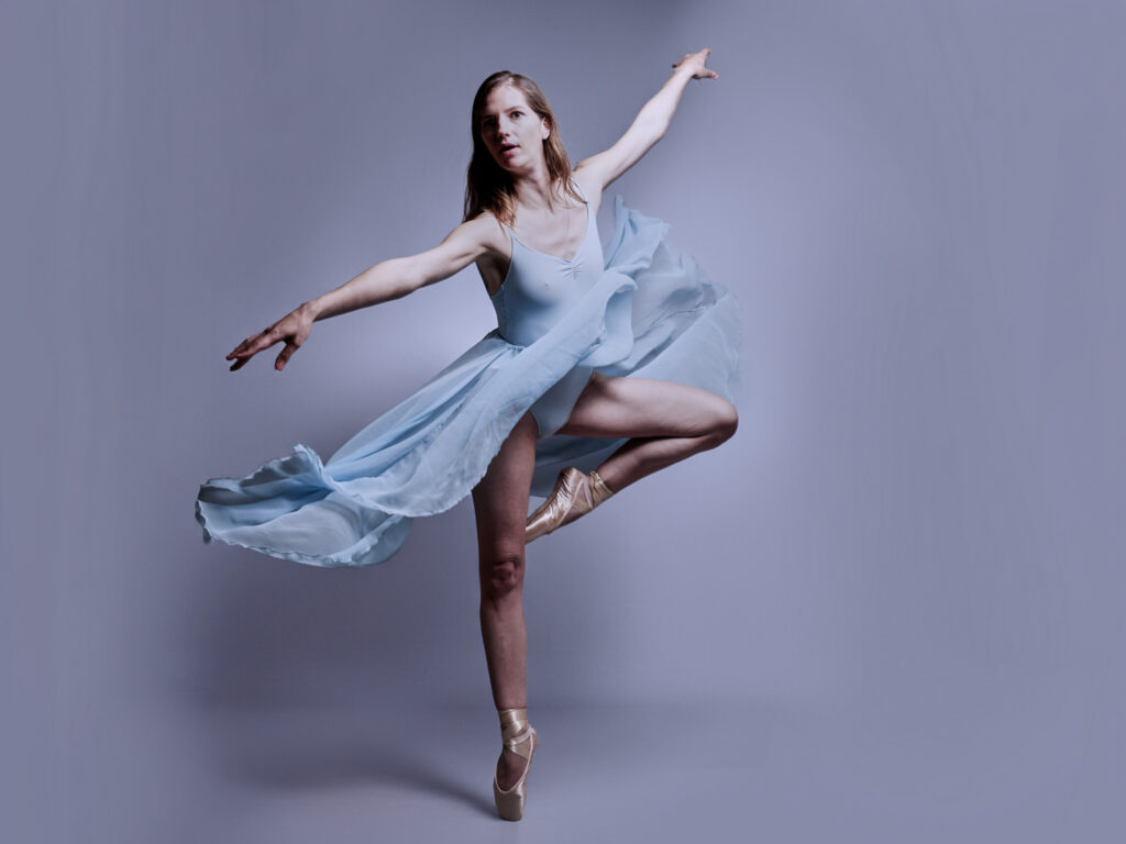 La danseuse Aurélie en équilibre sur une jambe, avec l'autre jambe levée. Elle est vêtue d'un justaucorps et d'une jupe bleu clair.