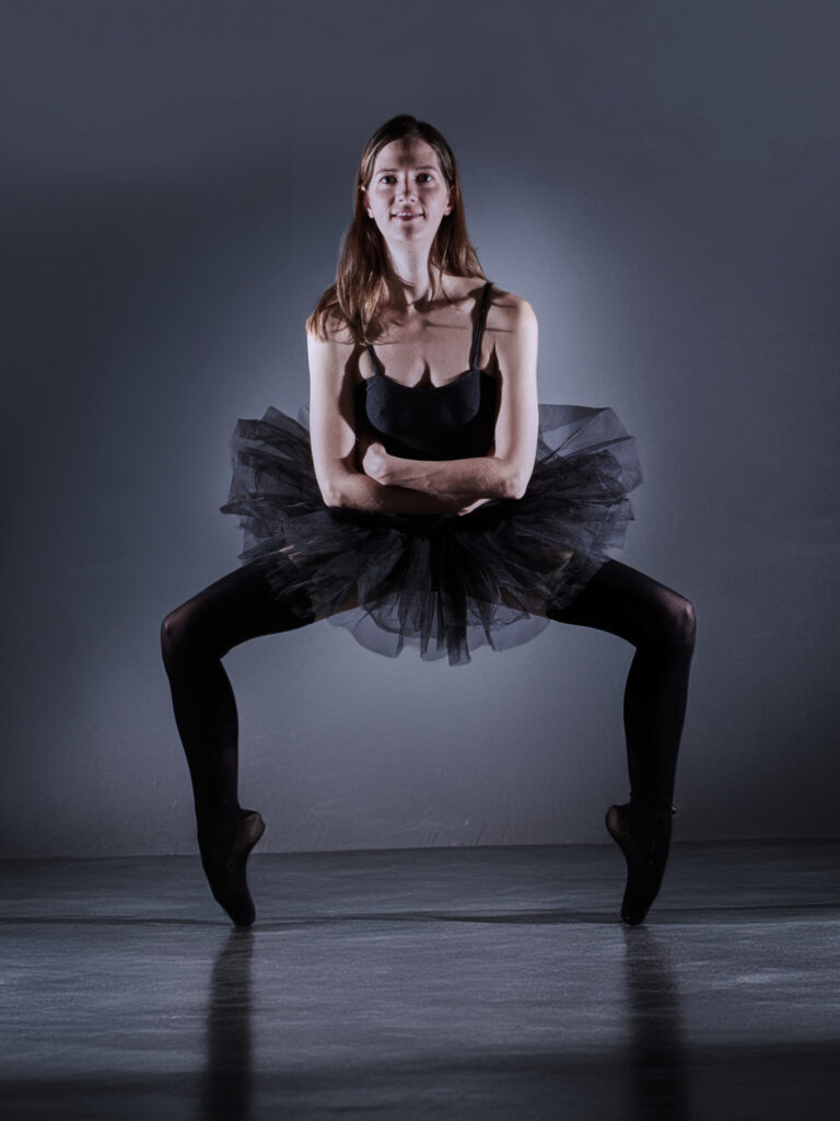 Aurélie, danseuse de ballet, est en position de plie, avec les bras croisés sur sa poitrine, vêtue d'un justaucorps et d'un tutu noirs. Elle a les pointes des pieds en dehors.