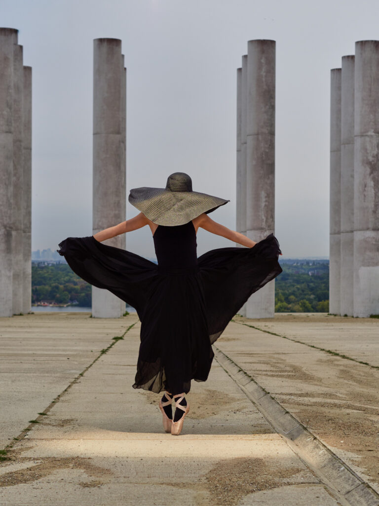 Photographie artistique d'une danseuse de dos en tutu et grand chapeau noir, sur pointes, devant des colonnes en béton. Jeu de symétrie et de perspective.