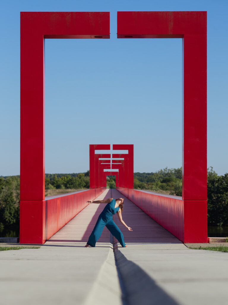 Danseuse en combinaison bleue sur un pont rouge vif. La pose en triangle est encadrée par des portiques rouges, créant une forte symétrie et une ligne de fuite.