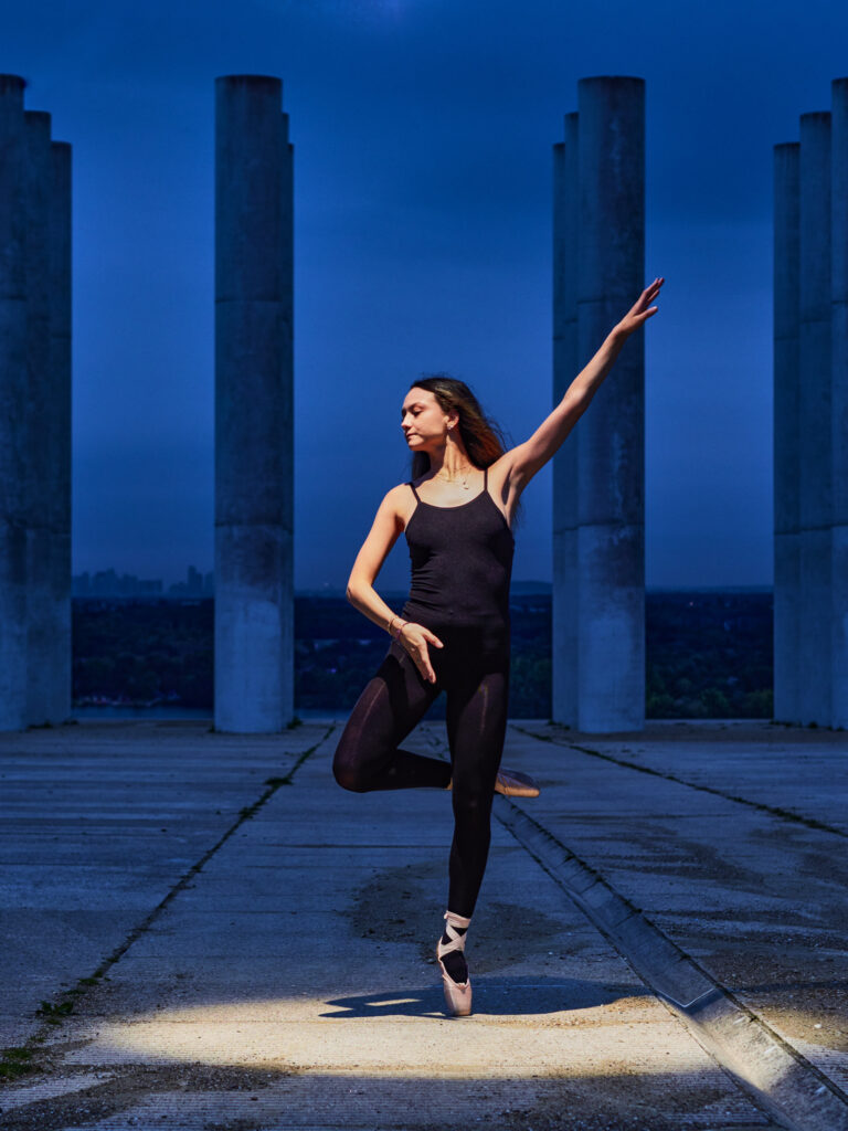 Danseuse en justaucorps noir sur une jambe, posant en équilibre sur pointe. Éclairage au sol focalisé sur le corps devant des colonnes de béton, ambiance nocturne bleue.
