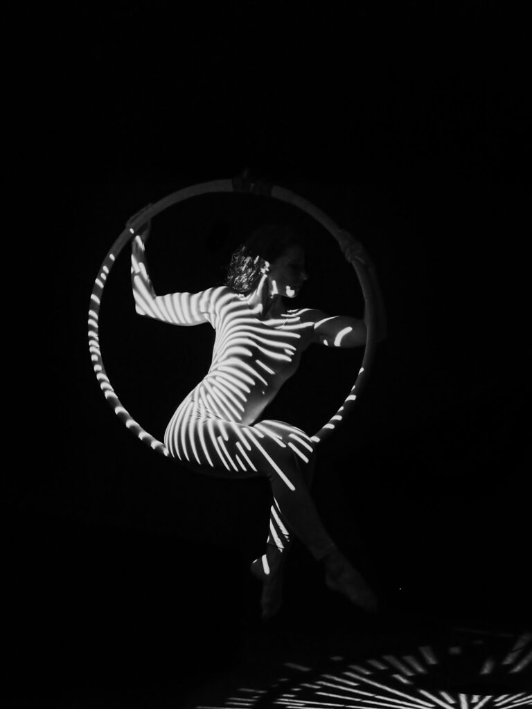Photographie noir et blanc d'une artiste suspendue sur un cerceau aérien (lyra), son corps éclairé par des rayures de lumière horizontale sur fond noir.