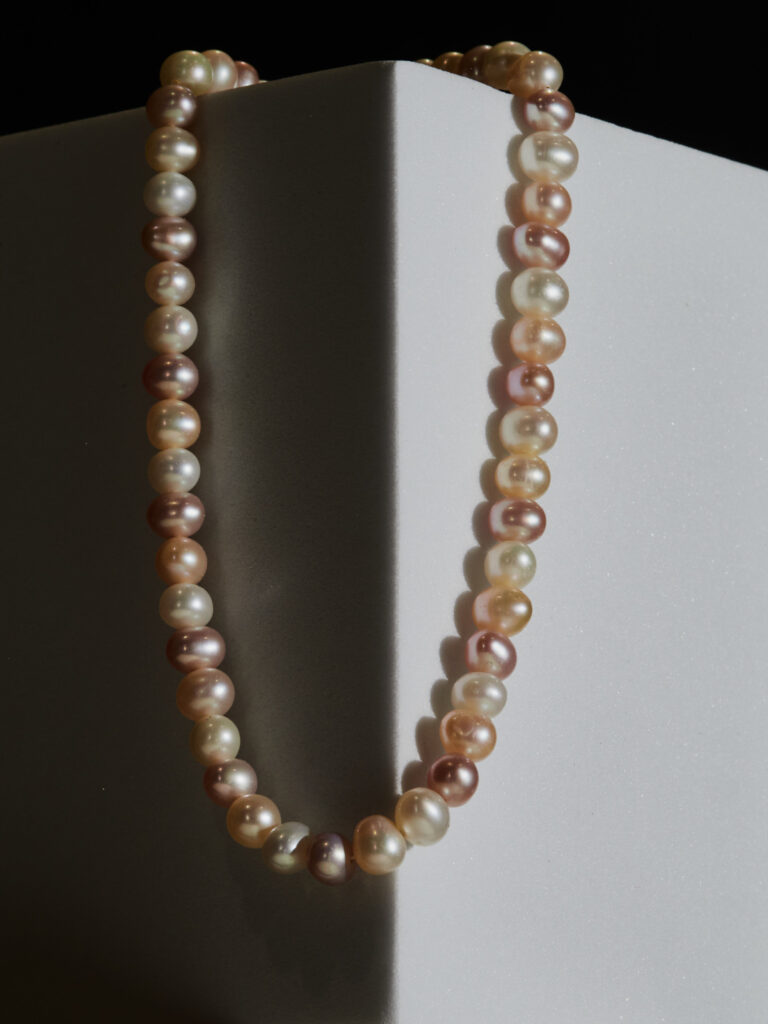 Collier de perles de culture aux teintes naturelles et rosées, photographié sur un fond géométrique blanc avec jeux d'ombres.