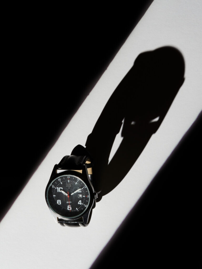 Montre-bracelet à cadran noir et bracelet en cuir, photographiée avec un éclairage de studio très contrasté projetant une longue ombre graphique.