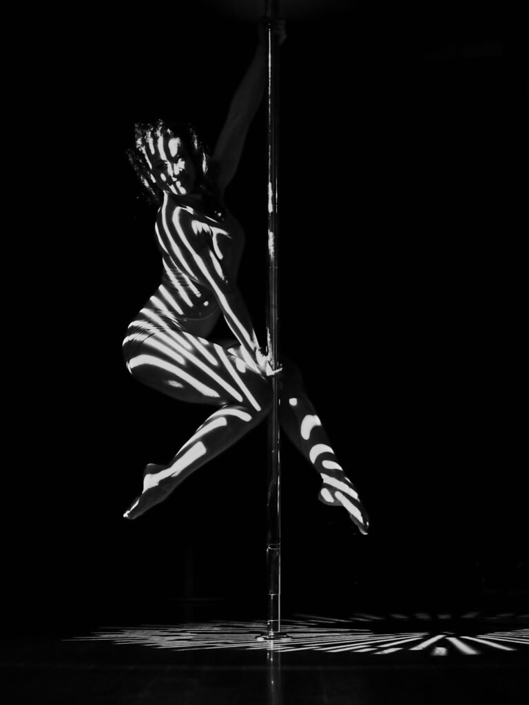 Photographie noir et blanc de pole dance artistique. Une acrobate est suspendue en l'air sur une barre verticale, son corps éclairé par des rayures de lumière blanche créant un effet de zèbre graphique sur fond noir.