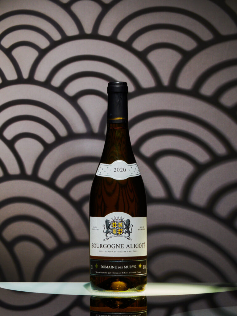 Photographie produit (packshot) de la bouteille de Bourgogne Aligoté Domaine des Murys (AOP). Arrière-plan graphique avec un motif de vagues japonisantes. Style luxe et lumière sculptée pour le e-commerce vin blanc.