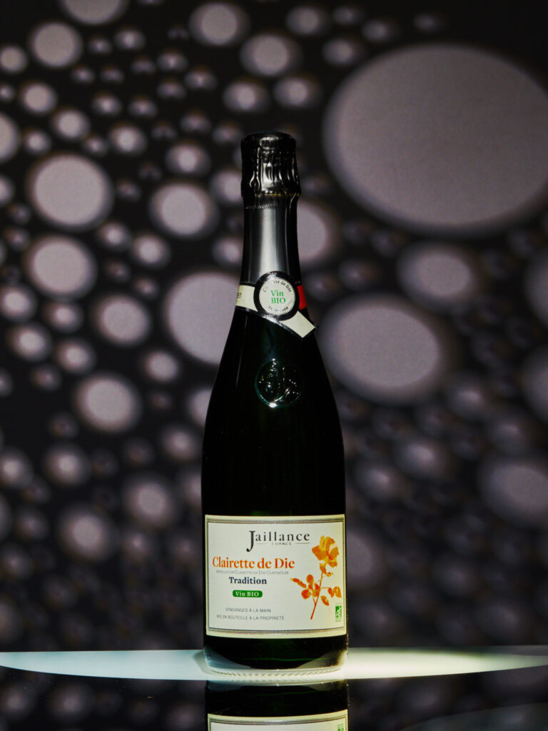 Photographie produit (packshot) de la bouteille de Clairette de Die Jaillance (Vin Bio, AOP), éclairage dramatique avec un motif de points lumineux en arrière-plan. Style luxe, lumière sculptée, idéal pour e-commerce.
