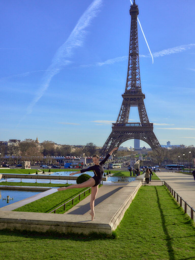 Lola en justaucorps noir effectue une arabesque parfaite sur pointes sur un muret des jardins du Trocadéro, face à la Tour Eiffel.