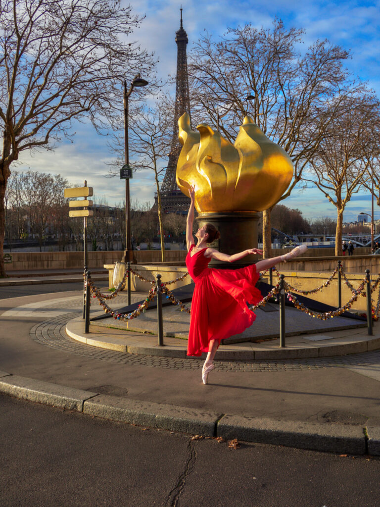 Daria (@daria_en_pointe) en pose de danse classique en robe rouge devant la sculpture dorée de la Flamme de la Liberté à Paris, avec la Tour Eiffel en arrière-plan.