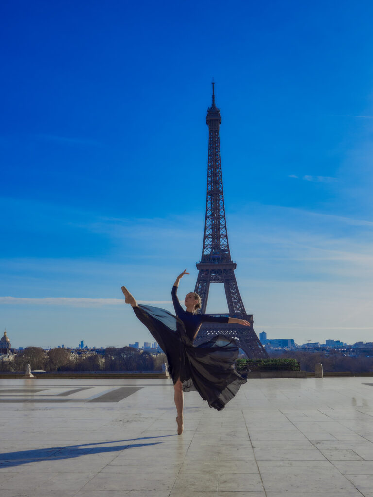 Portrait en pied de la danseuse Lola réalisant un développement à la seconde avec sa jupe noire fluide tourbillonnante sur l'esplanade du Trocadéro, face à la Tour Eiffel.
