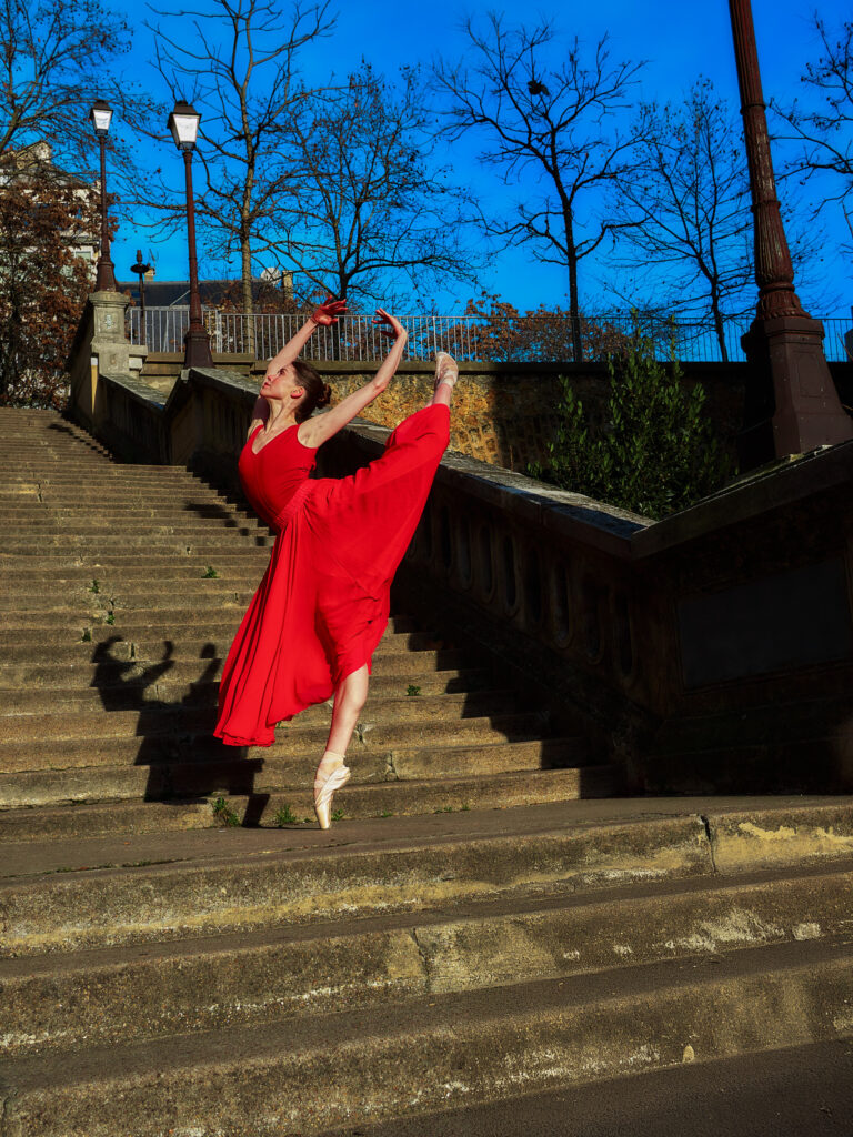 Daria en équilibre sur une pointe sur une marche d'escalier en pierre à Paris, réalisant un grand battement, sous un ciel bleu pur.