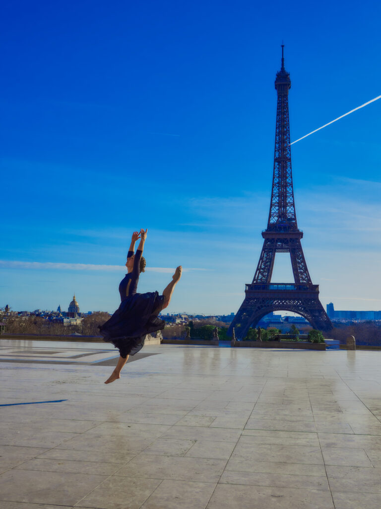 Lola (@lolite.danse) capturée en plein saut (grand jeté) au-dessus de l'esplanade du Trocadéro, avec la Tour Eiffel et un ciel bleu dégagé en arrière-plan.