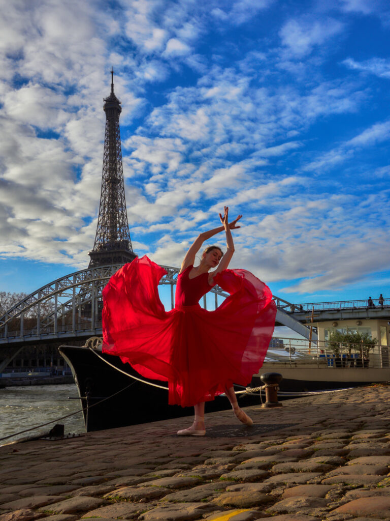 Portrait de la danseuse Daria en plein mouvement, sa robe rouge fluide volant au vent sur les pavés des quais de Seine, face à la Tour Eiffel et la passerelle Debilly.
