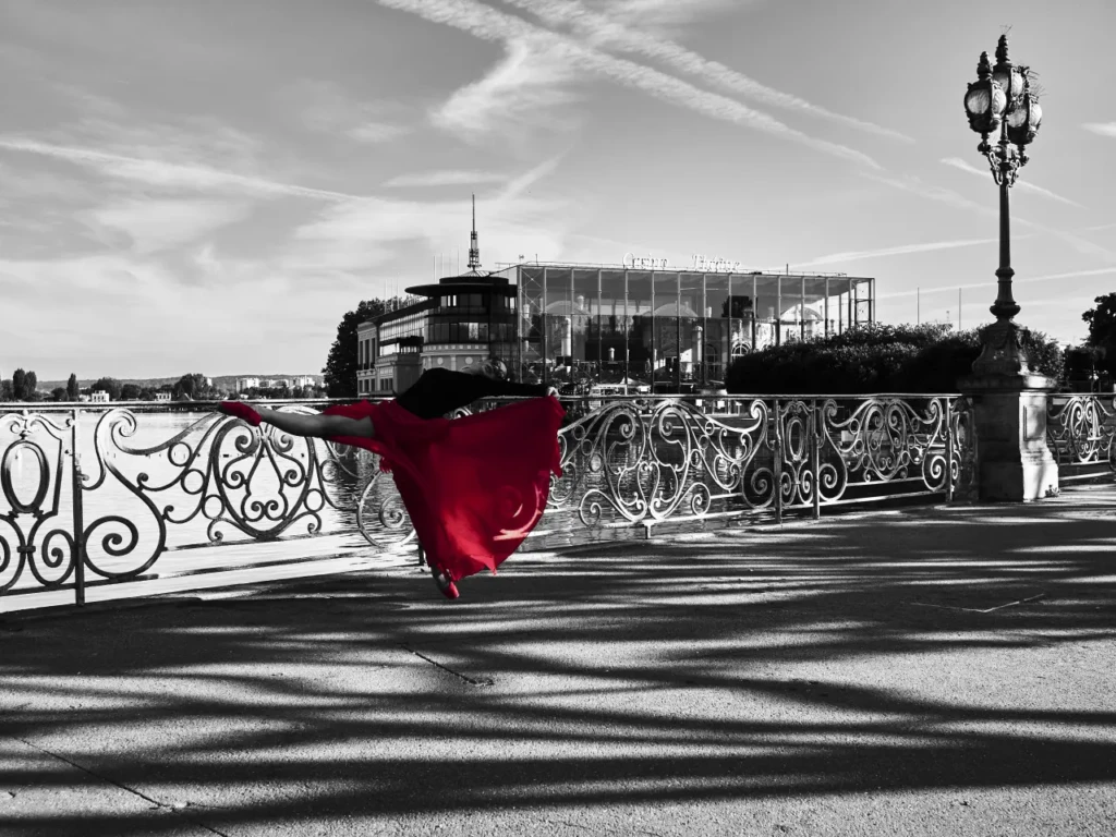 Danseuse en robe rouge réalisant une arabesque contre une rambarde, photo en noir et blanc avec couleur sélective.