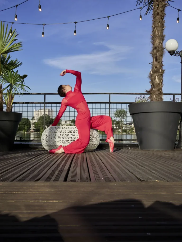 Danseuse en combinaison rouge agenouillée sur un pouf en osier blanc, terrasse d'Enghien-les-Bains.