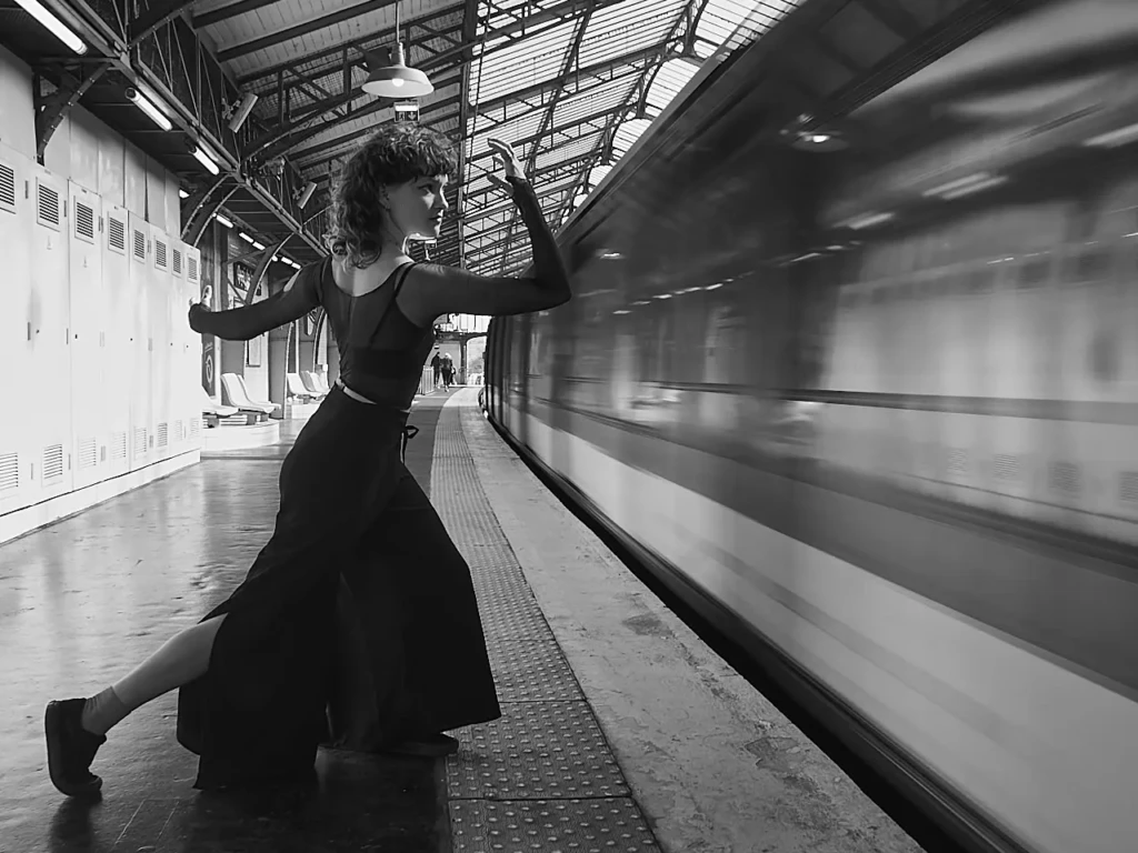 Danseuse en robe noire sur un quai de gare parisienne, train en mouvement flouté en arrière-plan.