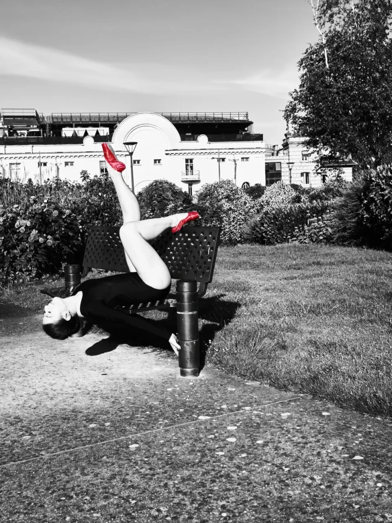 Danseuse en équilibre inversé sur un banc public, chaussons rouges en couleur sur fond noir et blanc.