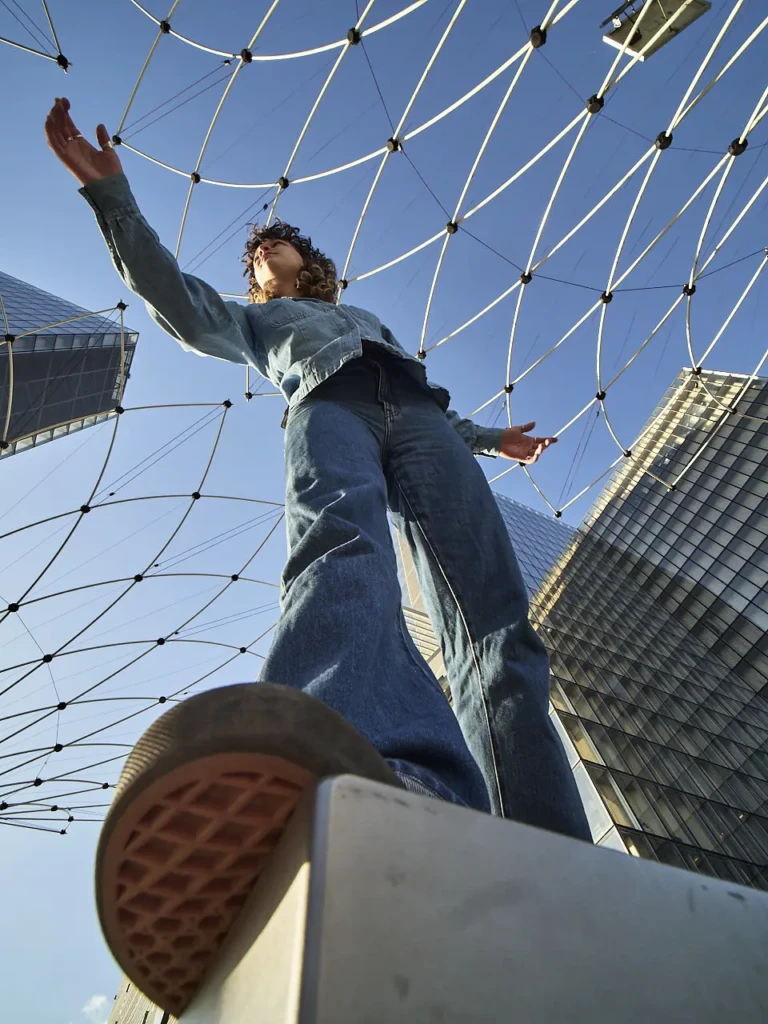 Danseuse en tenue denim debout, bras levés, sous une structure métallique en filet à la bibliothèque Francois Mitterrand.
