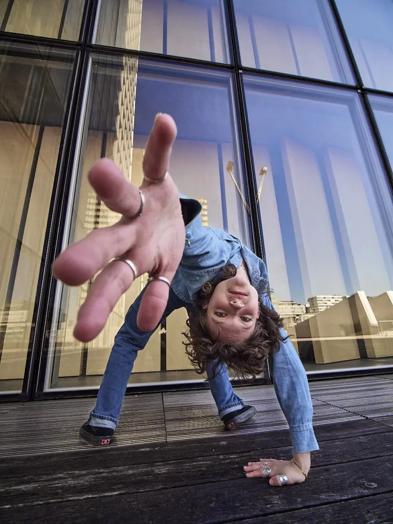 Danseuse en tenue denim tendant le bras vers l'objectif, photographiée en contre-plongée devant les verrières de la Bibliothèque François Mitterrand à Paris.