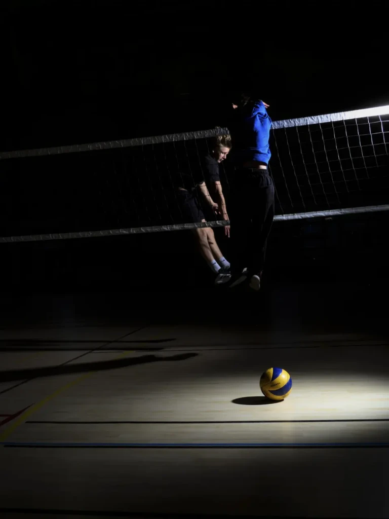 Deux joueurs de volley sautant ensemble pour un bloc au filet, ballon posé au sol éclairé.