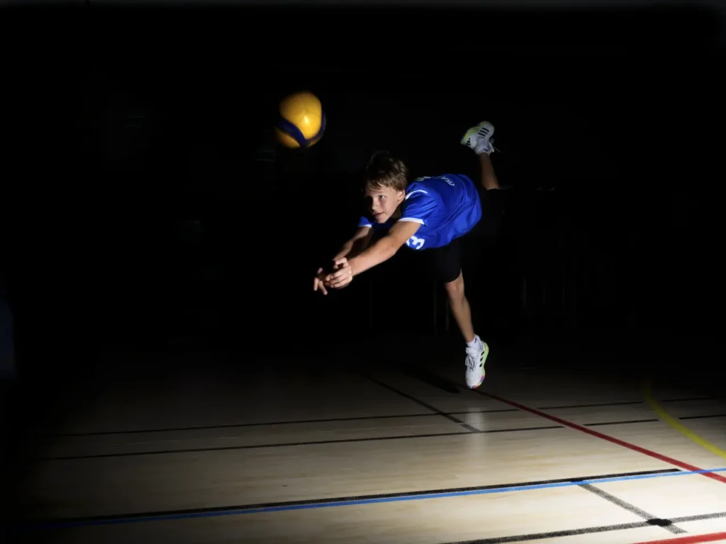 Joueur de volley en plongeon pour réceptionner le ballon, lumière sculptée sur fond noir.