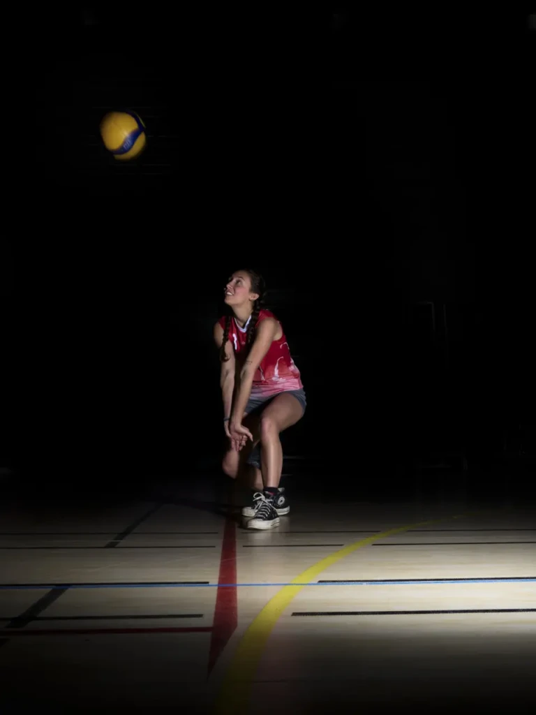 Joueuse de volley en position basse, suivant le ballon en plein vol, lignes de terrain éclairées.
